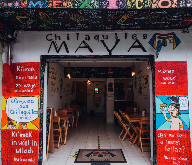 Exterior of Chilaques Maya restaurant storefront with red signage, wooden chairs visible inside, and colorful text banner above