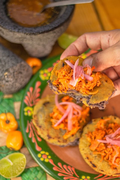 Hand holding a crispy taco topped with shredded carrot and pink onion, served on green plate with lime